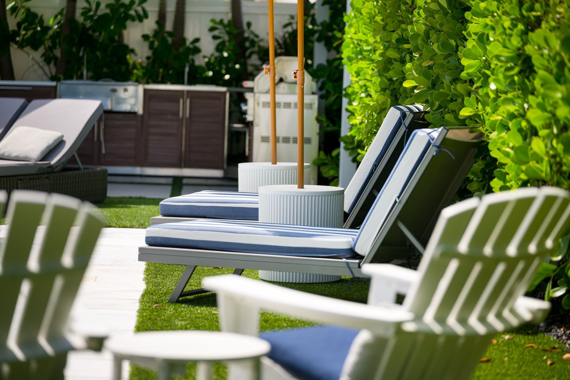 Outdoor lounge chairs and cushions on a sunny patio beside a green hedge.