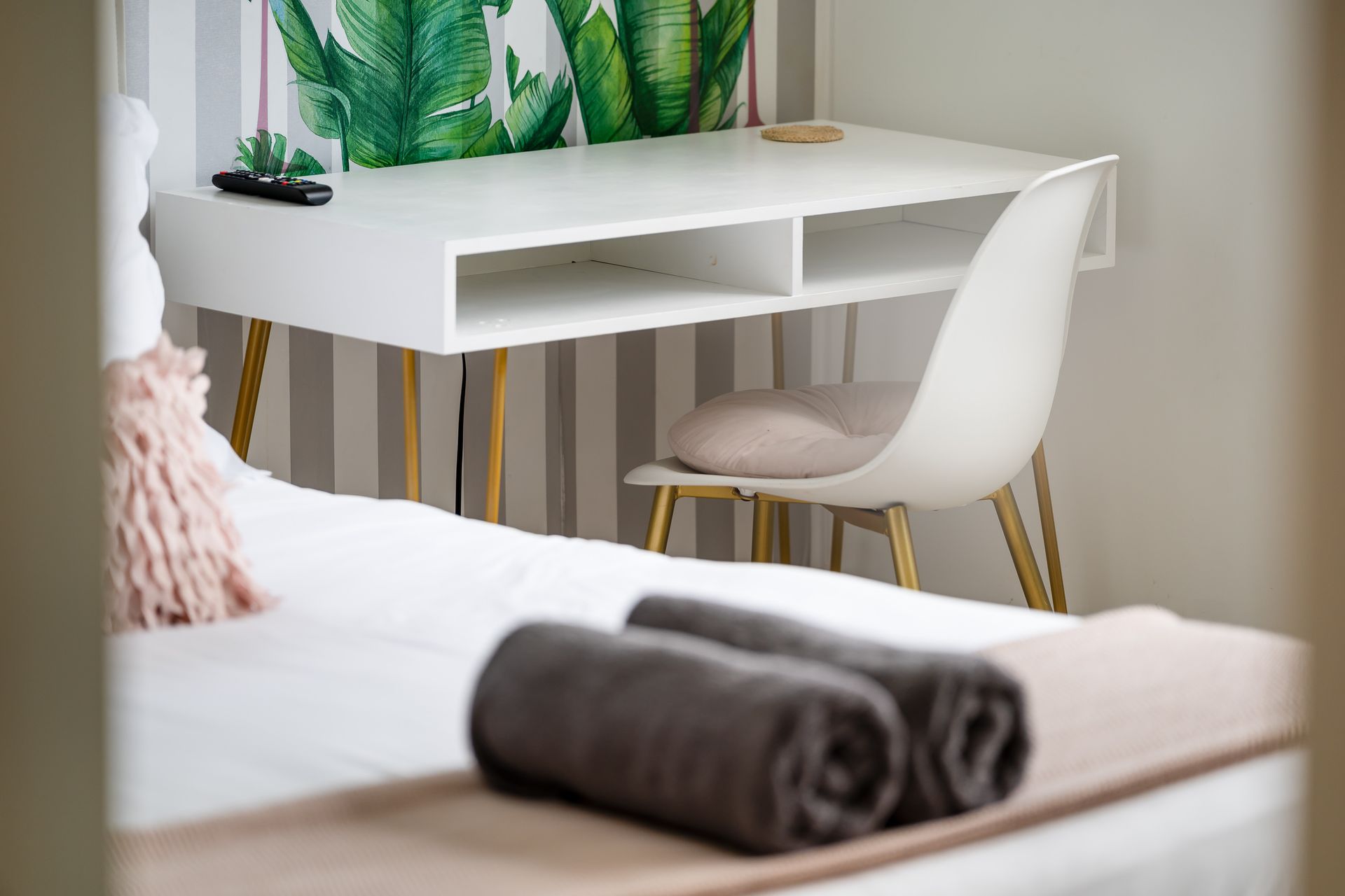 White desk with plant decor and white chair beside a bed with folded towels in a bright room