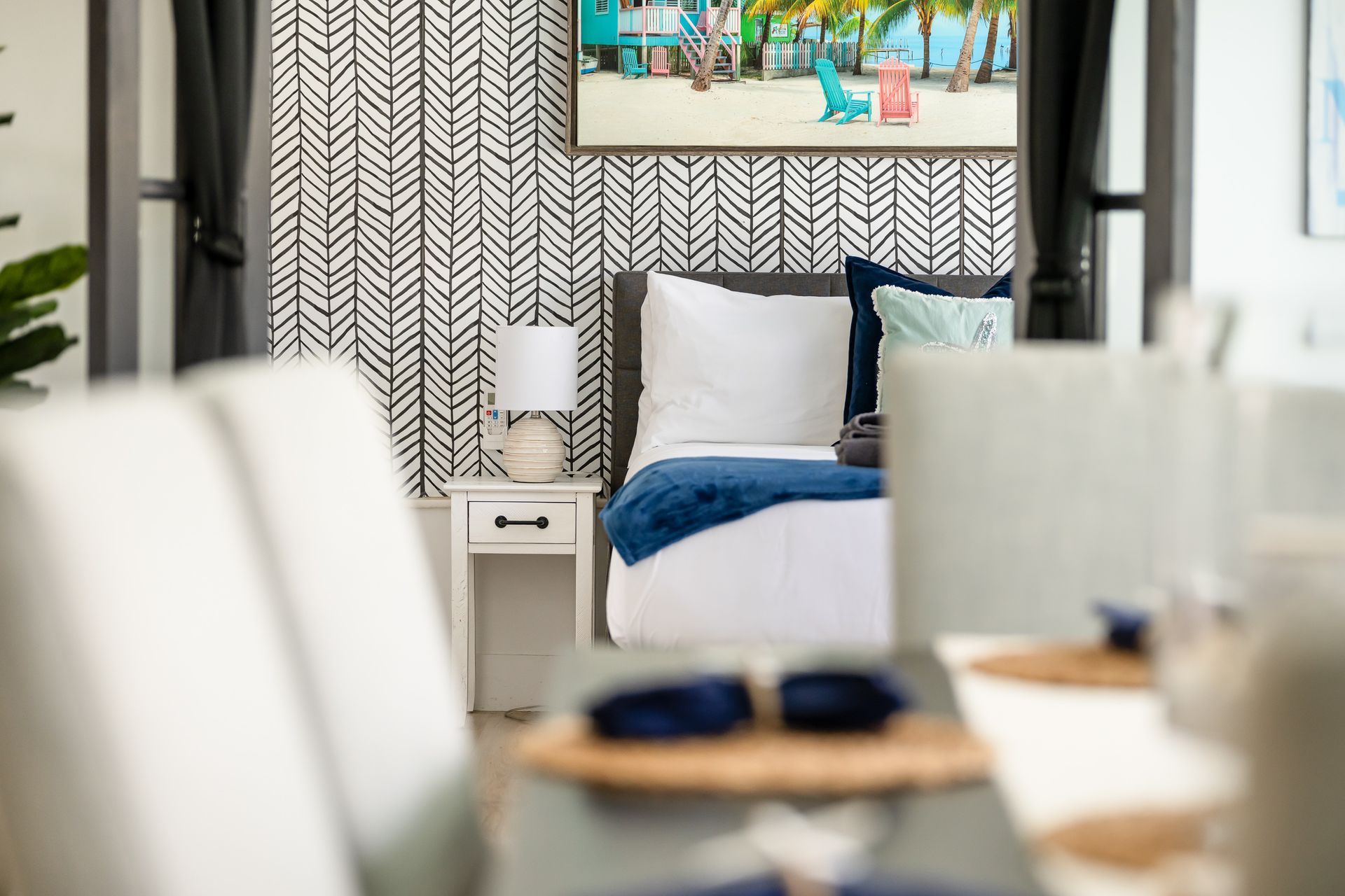 Cozy bedroom with a neatly made bed, patterned wallpaper, and a beach-themed painting above the headboard