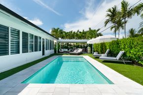 Modern backyard pool with white lounge chairs, palm trees, and a white house exterior