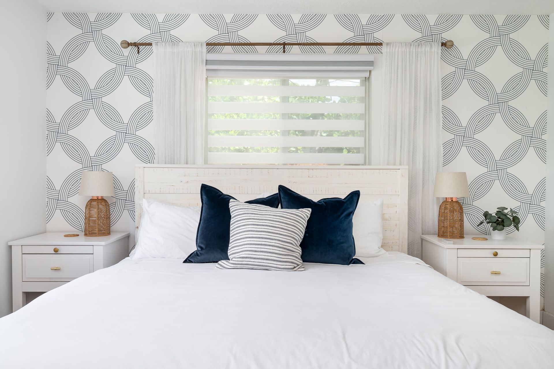 Bright white bedroom with patterned wallpaper, a large bed, and navy accent pillows beneath a window.