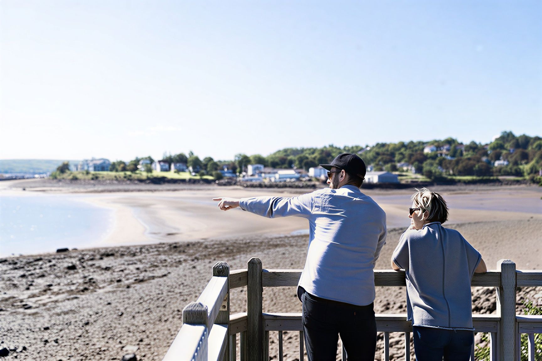 Two people stand on a boardwalk, pointing across a calm beach and bay toward houses on the opposite shore.