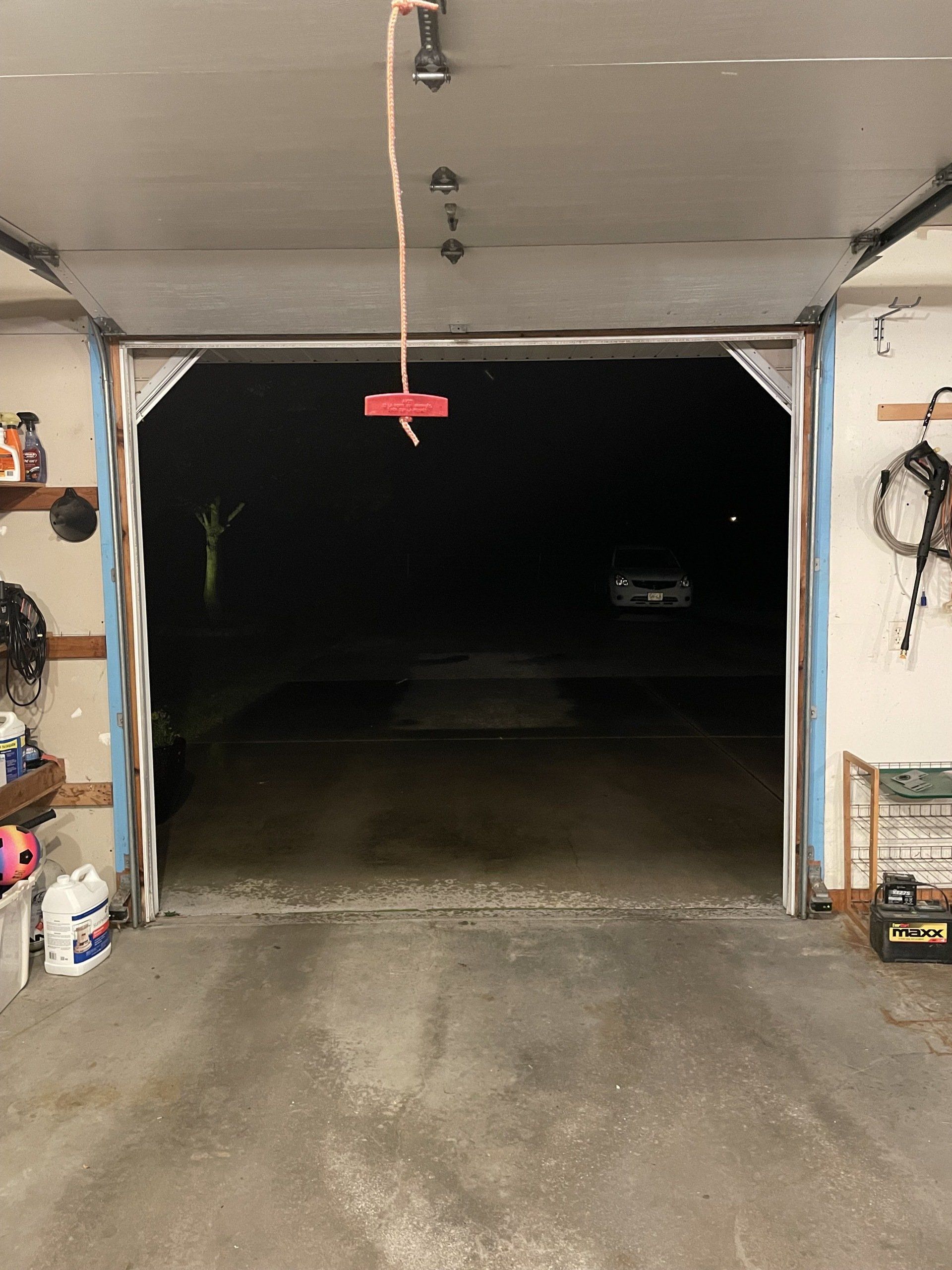Open garage door at night, with a concrete driveway and walls lined with tools and storage items.