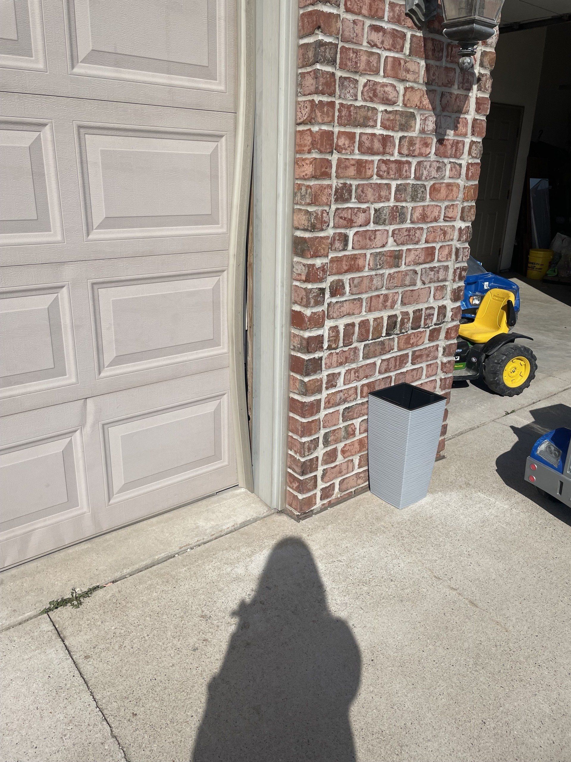 Brick house corner beside garage door, with trash bin, concrete driveway, and a yellow toy tractor in the background.