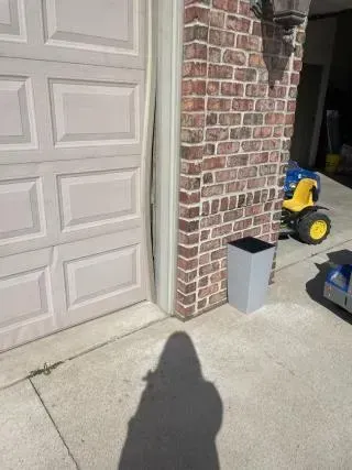 Brick house corner beside garage door, with trash bin, concrete driveway, and a yellow toy tractor in the background.