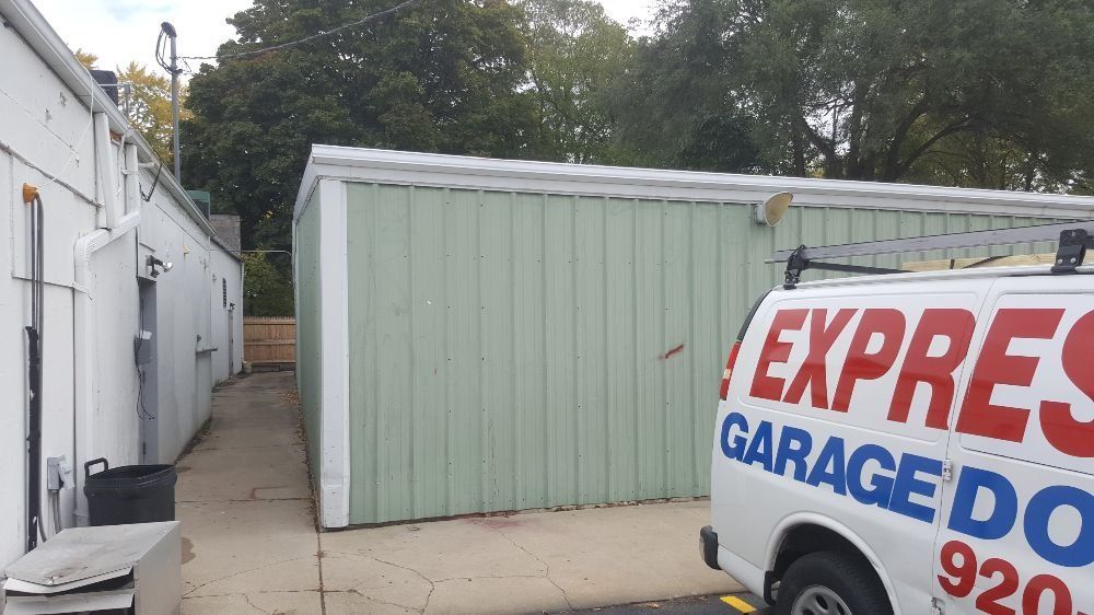 White industrial buildings beside a pale green garage; partial white service van with red and blue lettering in foreground