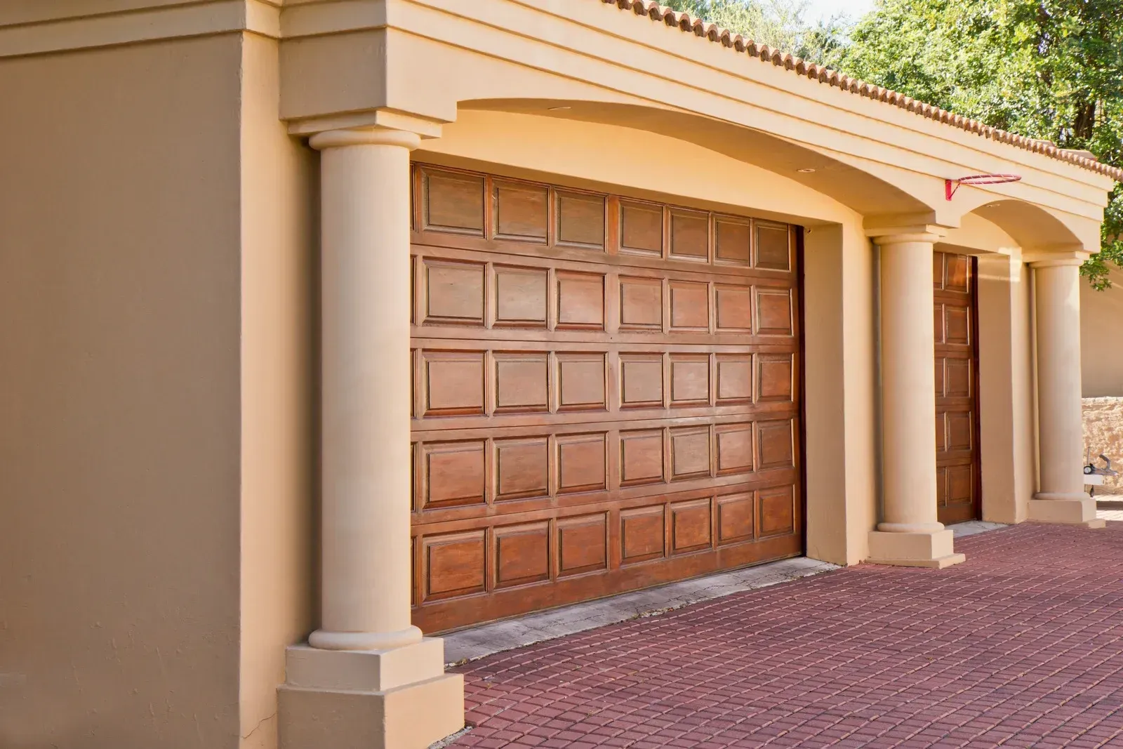 Tan garage with brown wooden door and columns, on a red brick driveway.