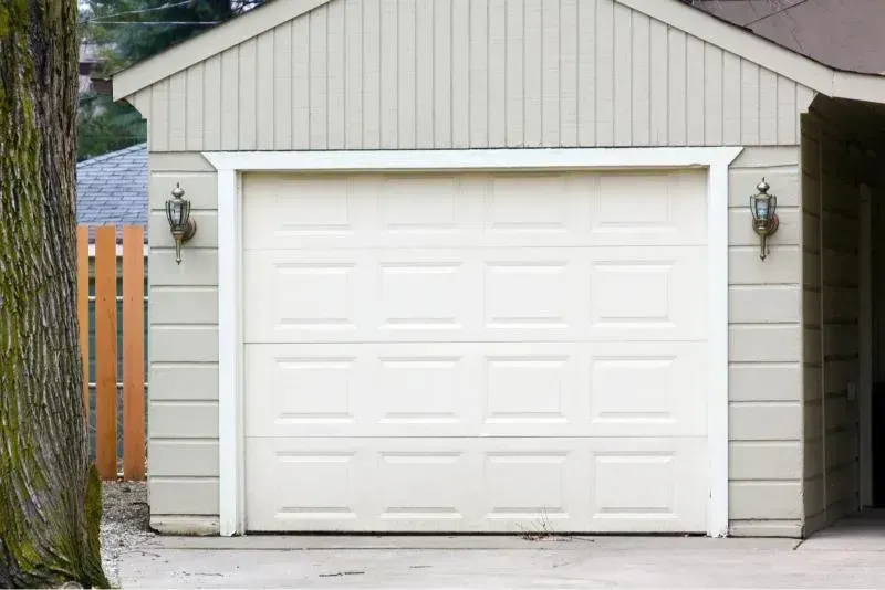White garage door on a gray detached garage with wall lanterns and a wooden fence beside it
