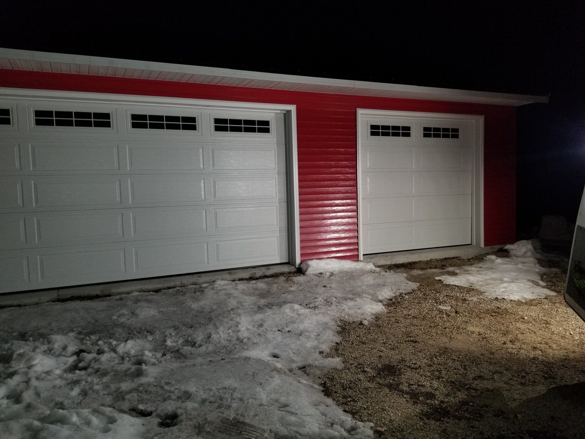 Snowy driveway in front of red house with two white garage doors at night