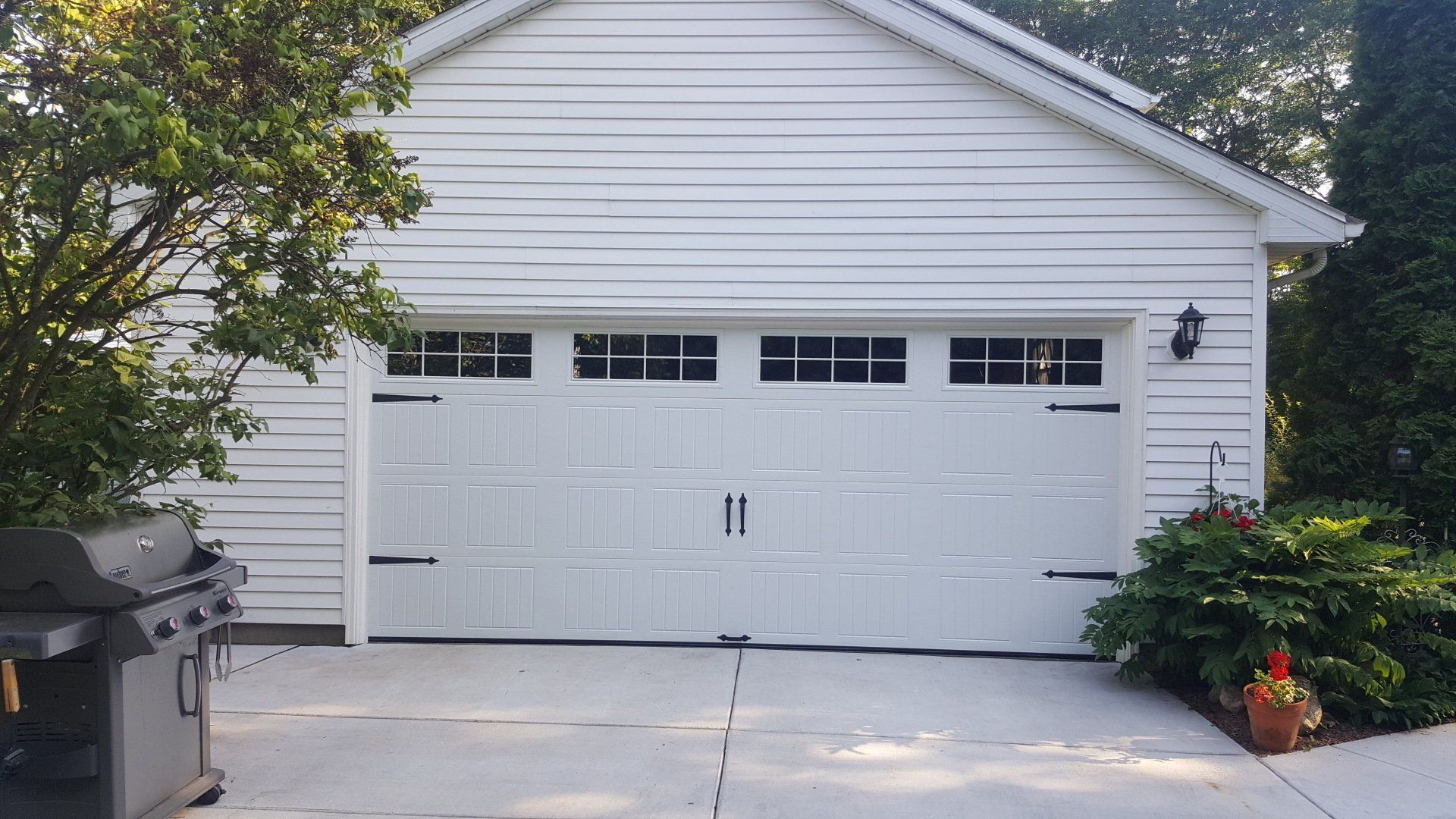 White two-car garage with driveway, surrounded by greenery and flowering plants.