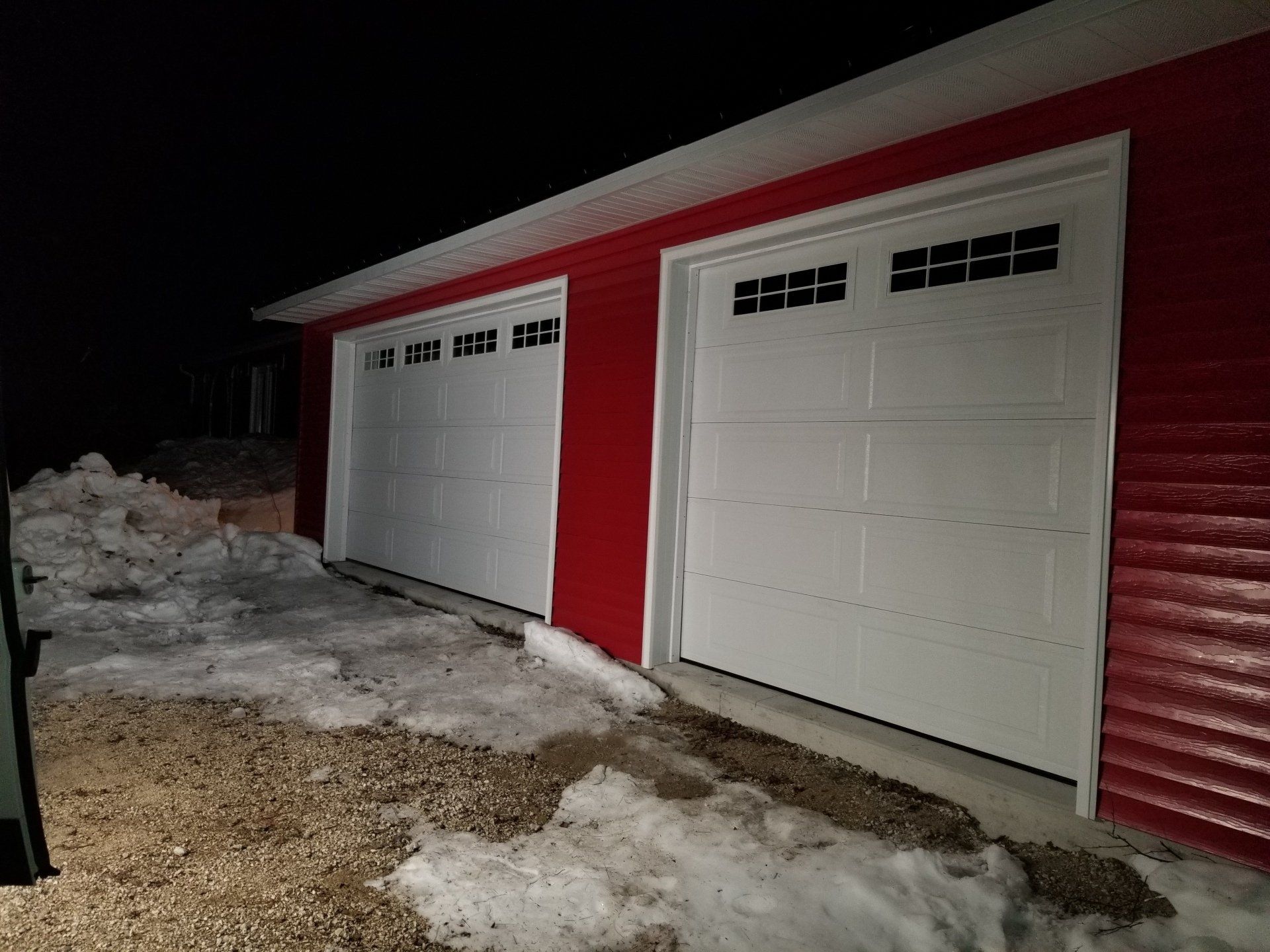 Red garage with two white doors beside a snow-covered driveway at night