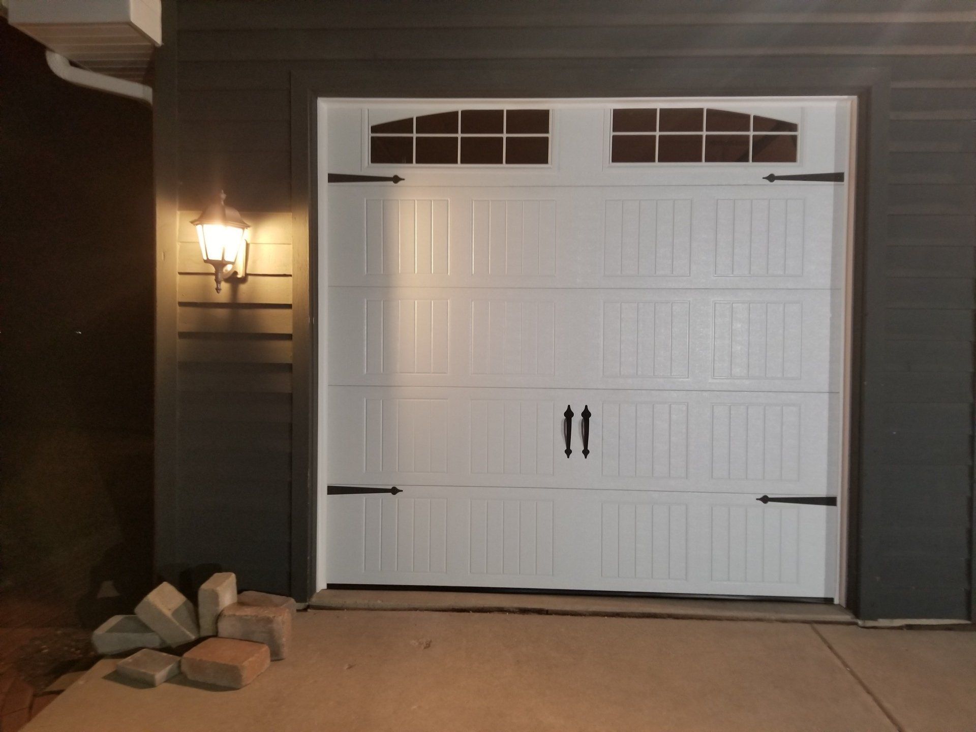 White garage door on a house exterior at night, lit by a porch light, with rocks beside the entrance.