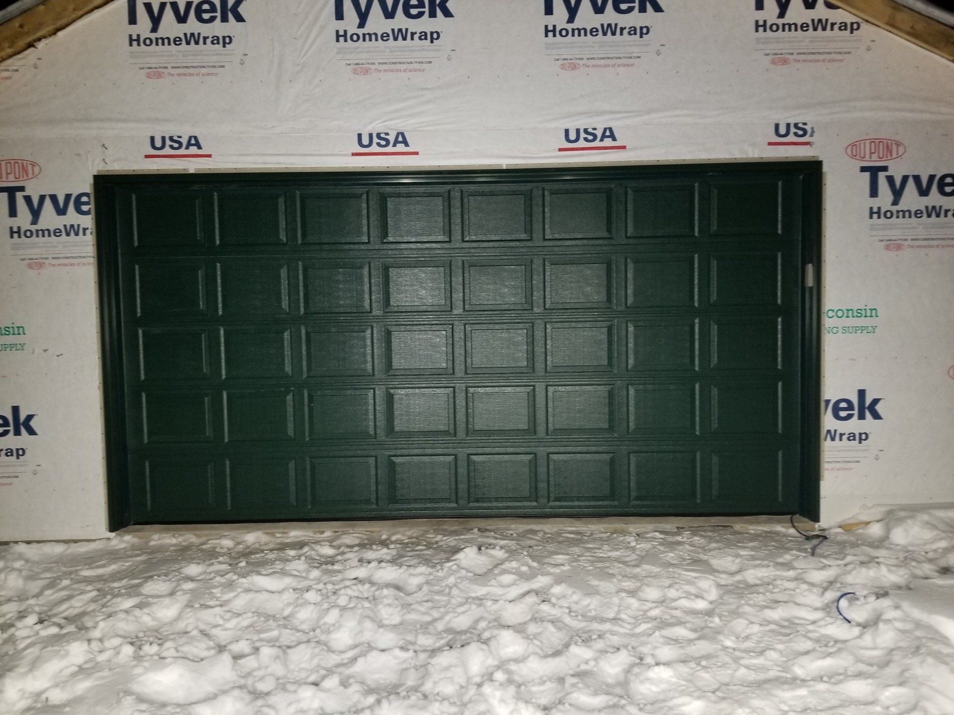 Dark green garage door on a house under construction, surrounded by Tyvek house wrap and snow.