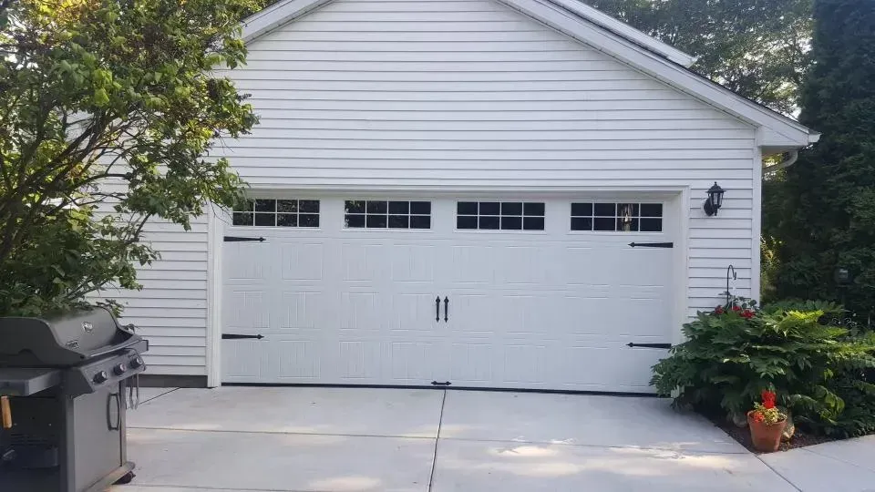 White two-car garage with driveway, surrounded by greenery and flowering plants.