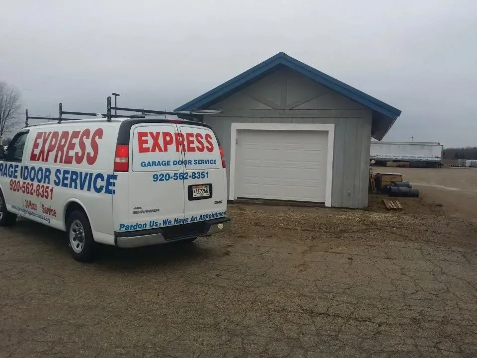 White Express van parked beside a small gray building with a blue roof on a gravel lot.