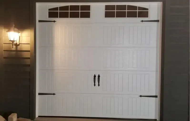White garage door on a dark house exterior with a lit wall sconce on the left