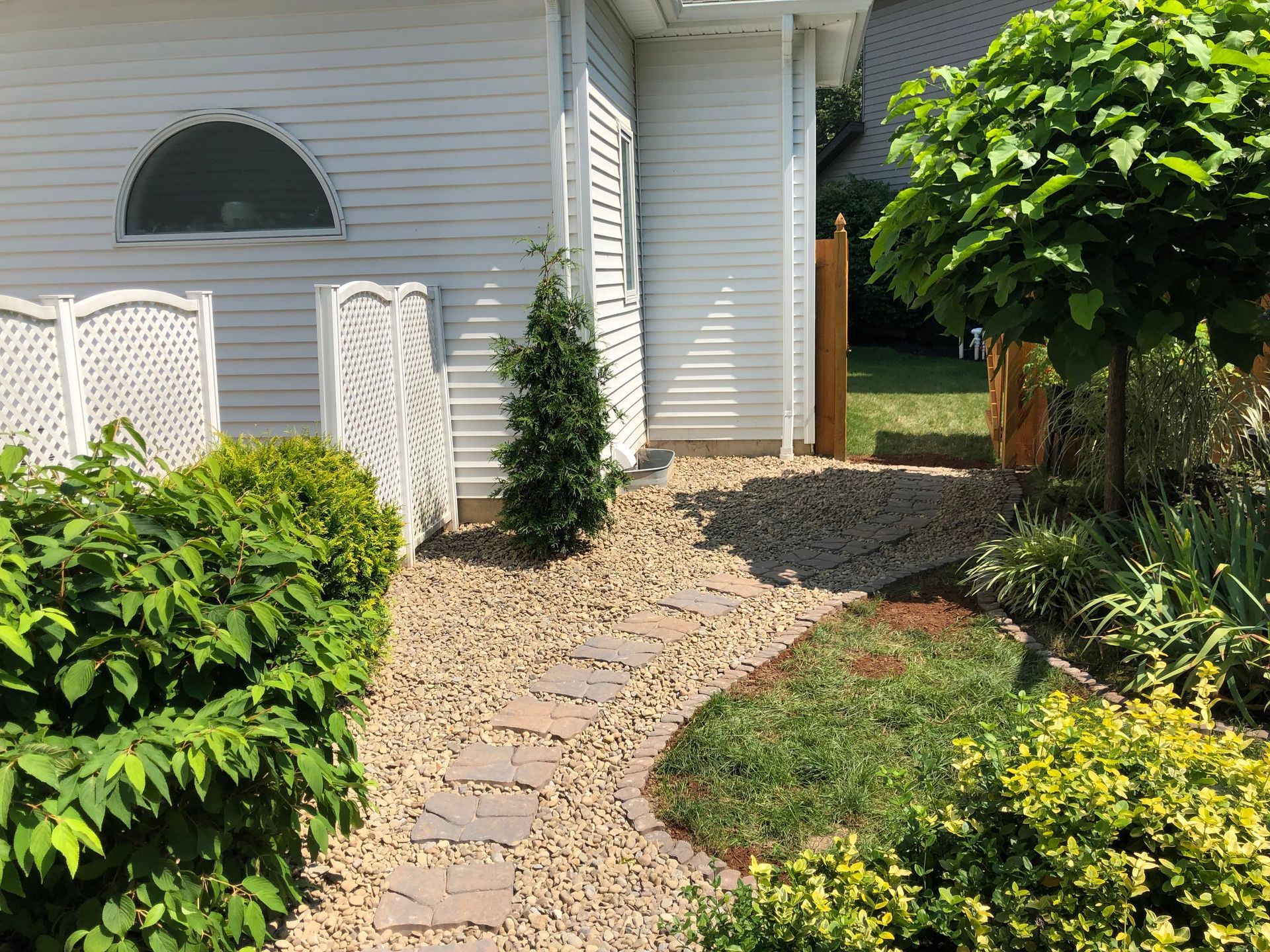 A stone pathway winds through a gravel yard beside a white-sided house, surrounded by lush green shrubs and a small tree.