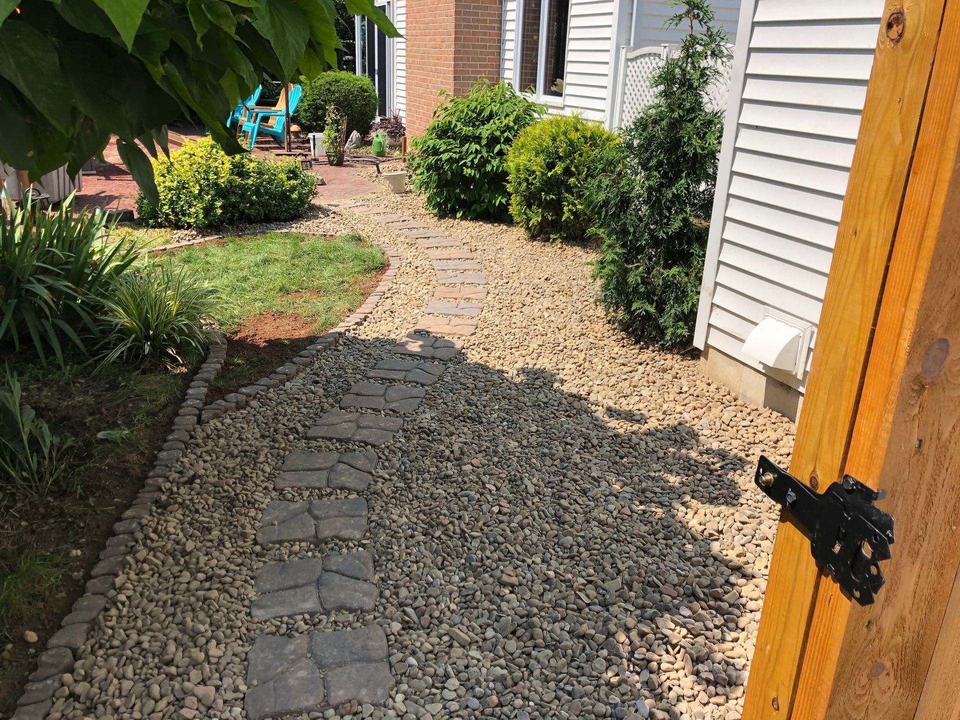 A stone paver walkway curves through a gravel yard leading to the side of a house with shrubs and a wooden fence post.