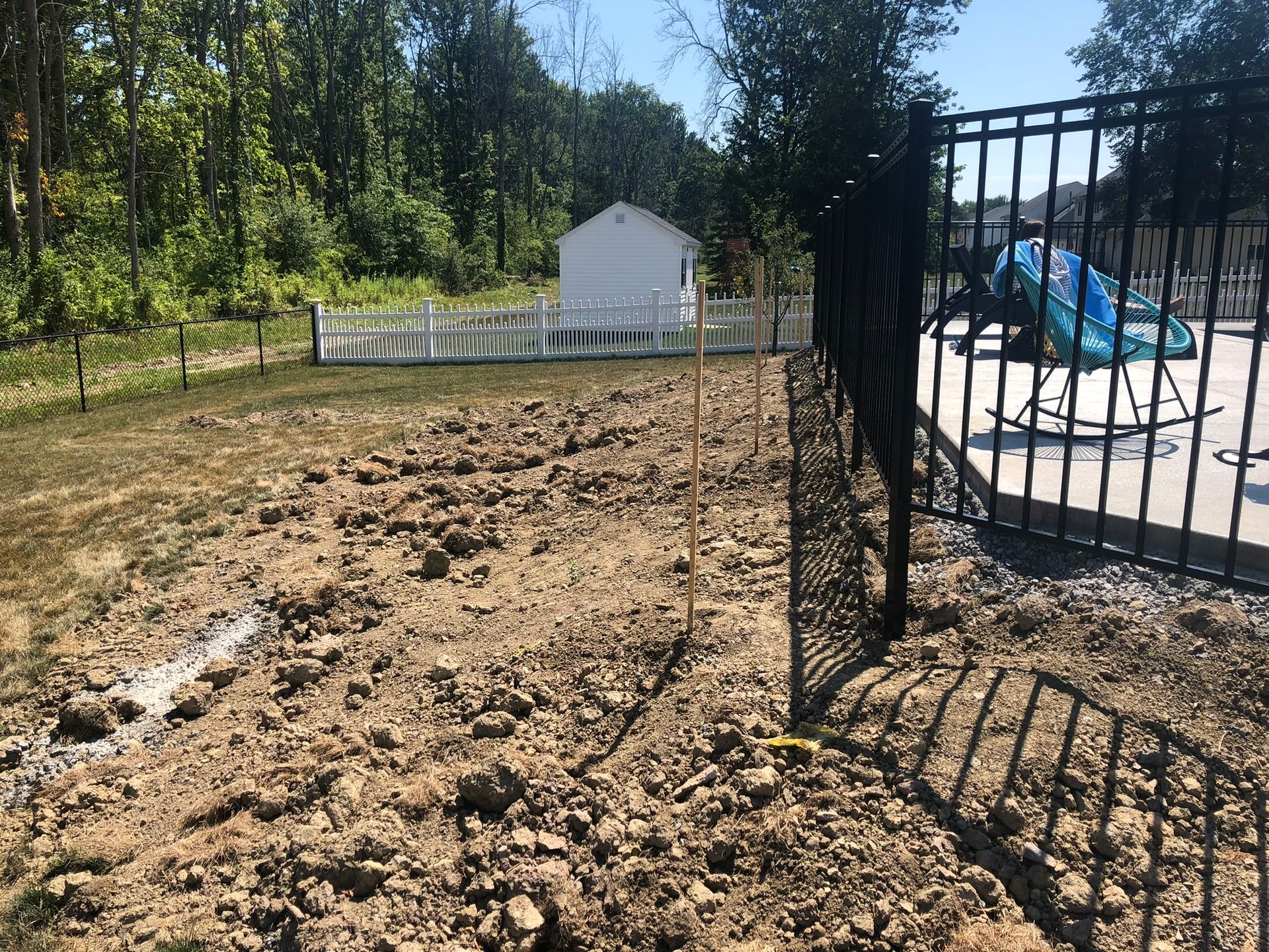 A dirt yard under construction next to a black metal pool fence and a small white shed in the distance.