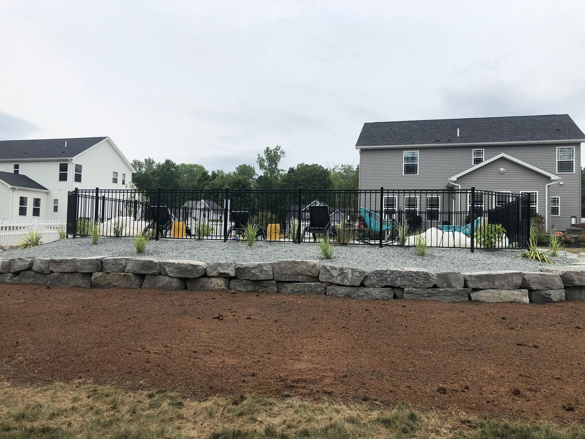 A retaining wall of grey stone blocks sits in front of a fenced pool area between two suburban houses.