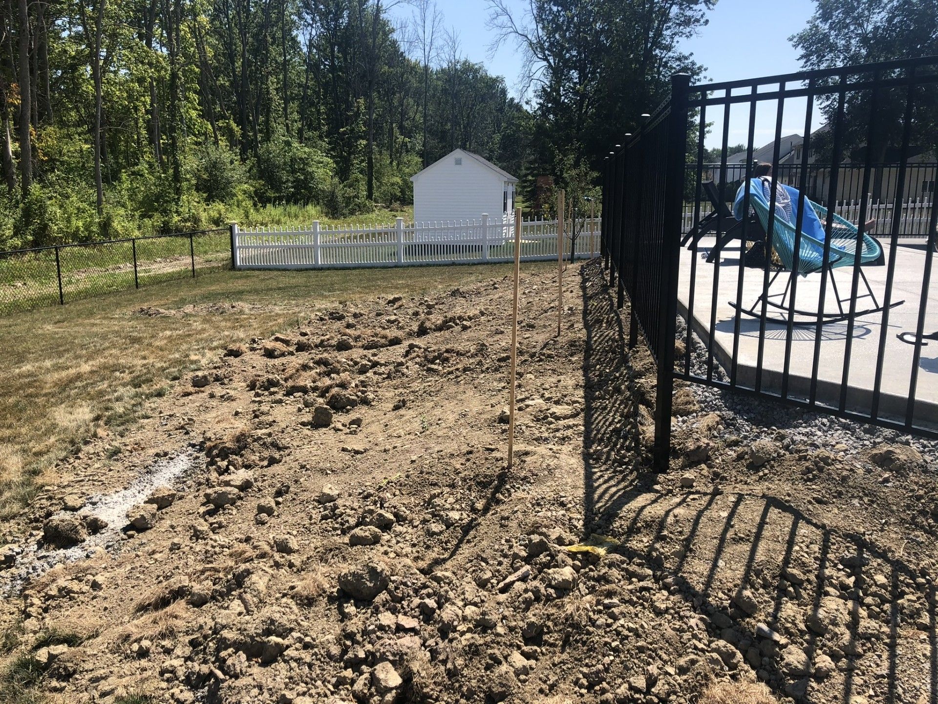 An uneven dirt yard with wooden stakes sits beside a black metal pool fence, with a small white shed in the background.