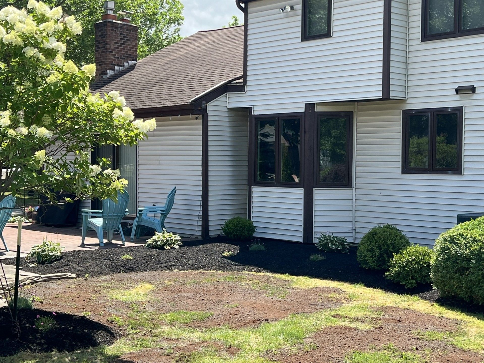 White house with dark window frames and a chimney, featuring a landscaped garden with shrubs, mulch, and blue patio chairs.