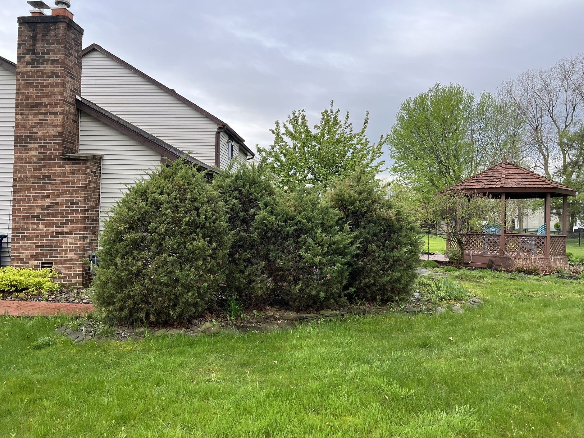 A brick chimney attached to a light-colored house next to a row of shrubs and a wooden gazebo in a grassy yard.