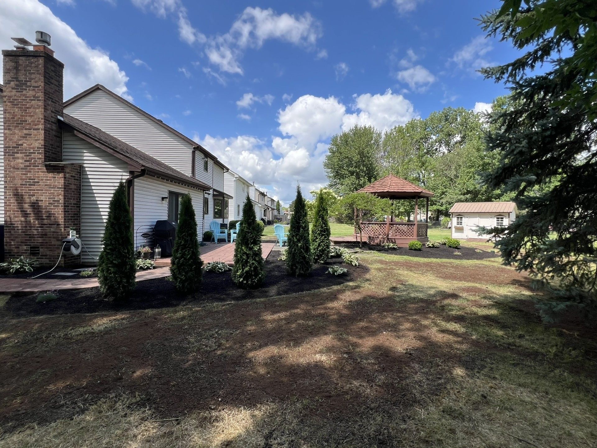 A brick and siding house with a patio, evergreen shrubs, a backyard gazebo, and a small shed under a blue, cloudy sky.