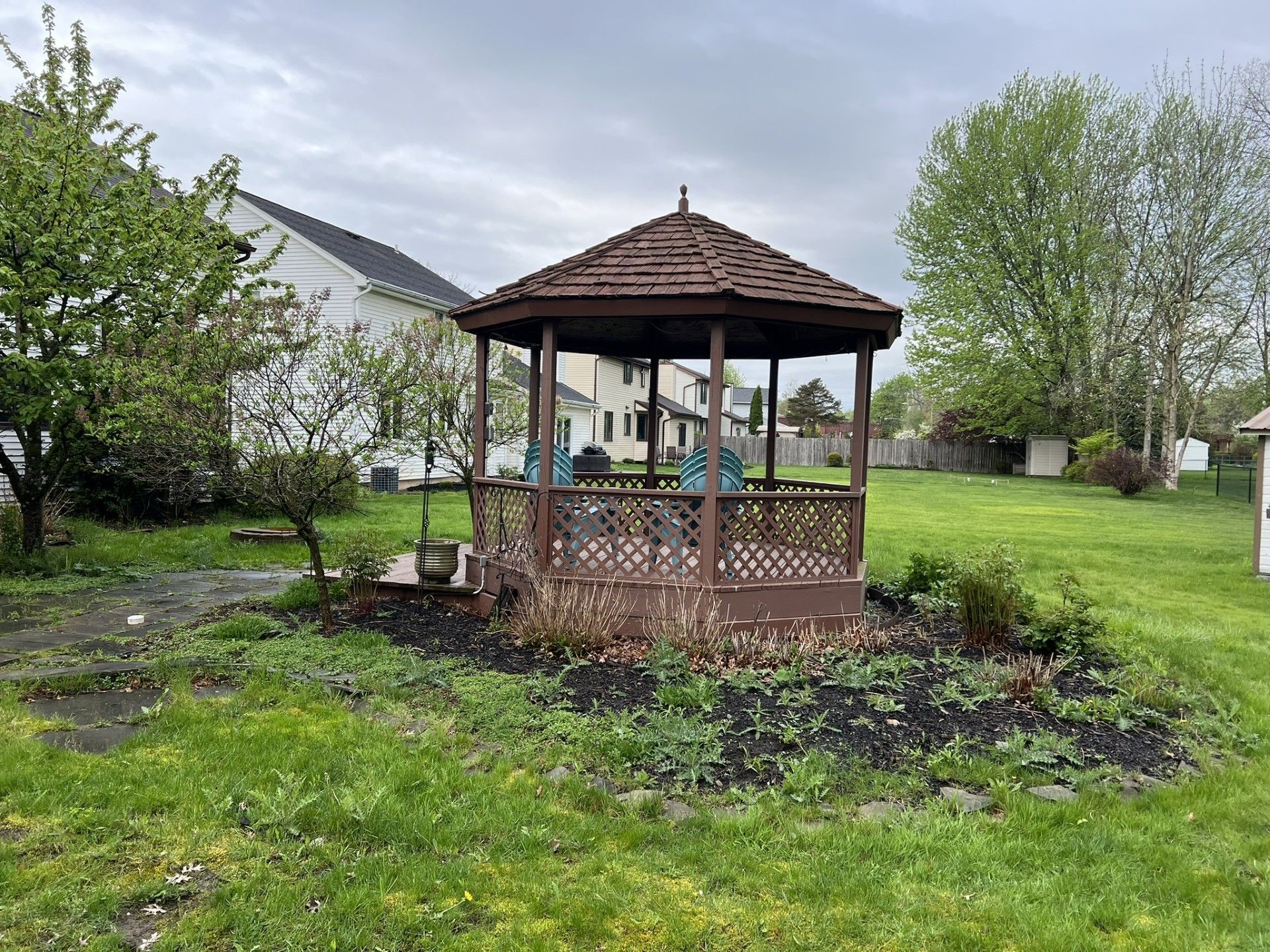 A brown wooden gazebo sits in a backyard surrounded by mulch, green grass, trees, and nearby houses under a cloudy sky.