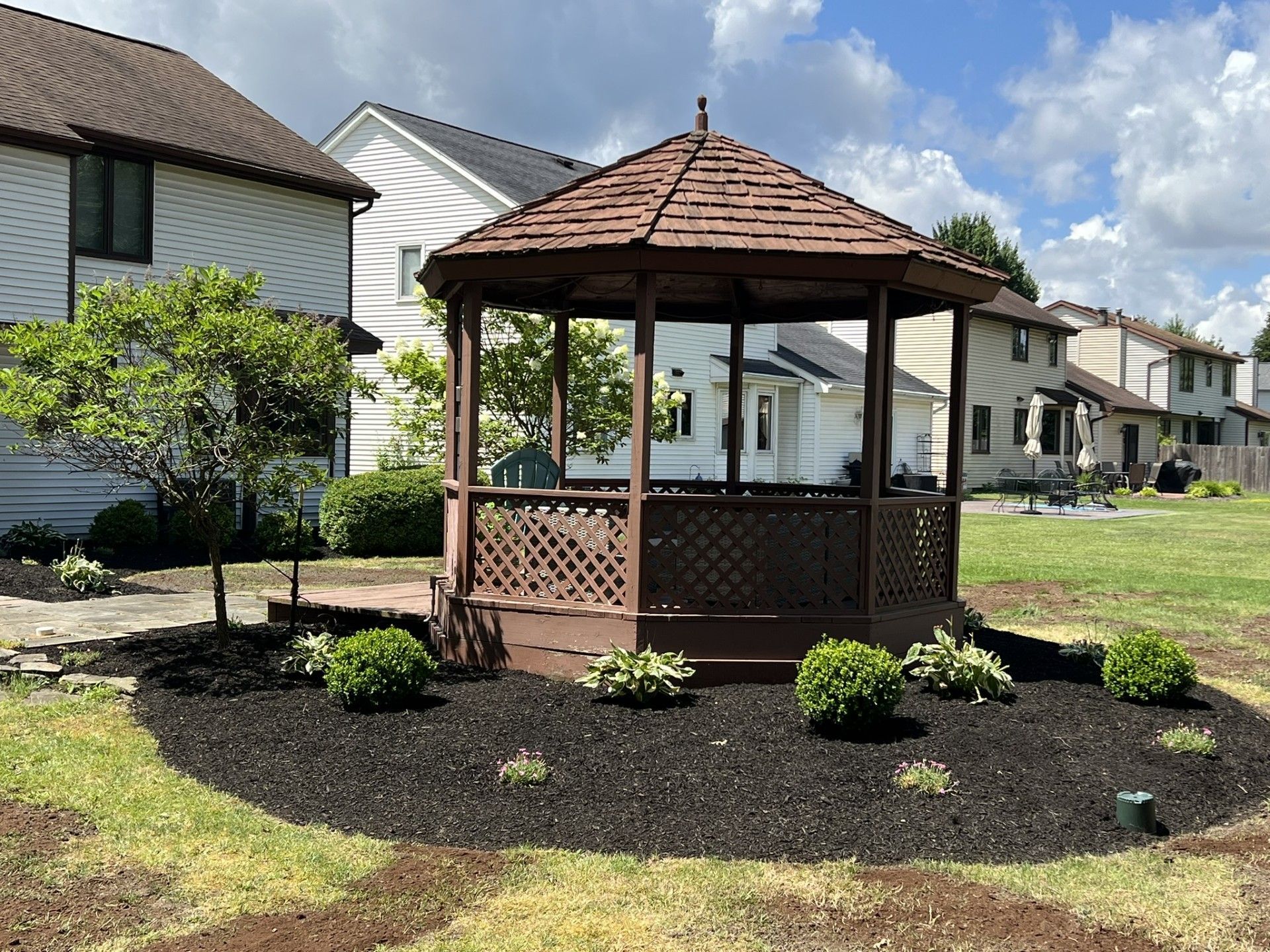 A brown wooden gazebo sits in a landscaped garden bed with mulch and small shrubs in a suburban backyard.