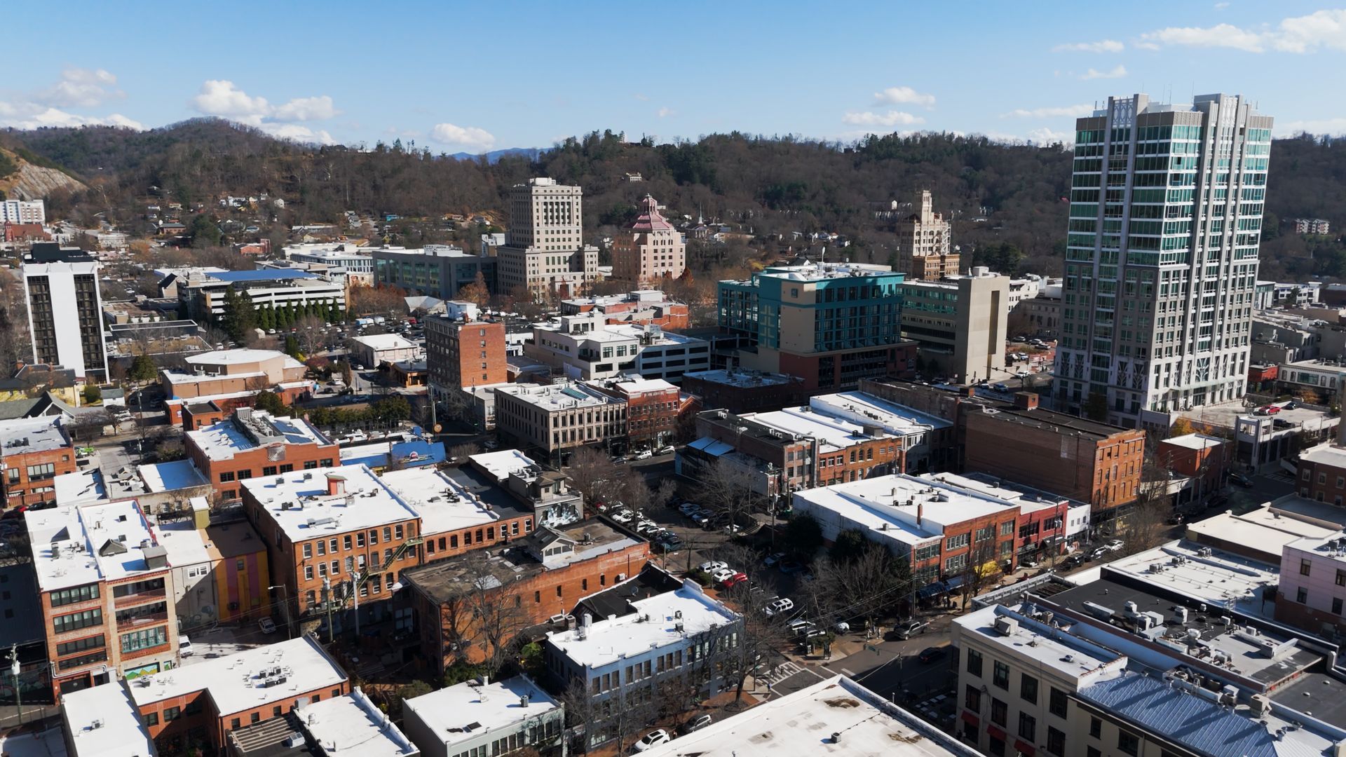 Snowy downtown cityscape with brick buildings, a tall glass tower, and hills in the background