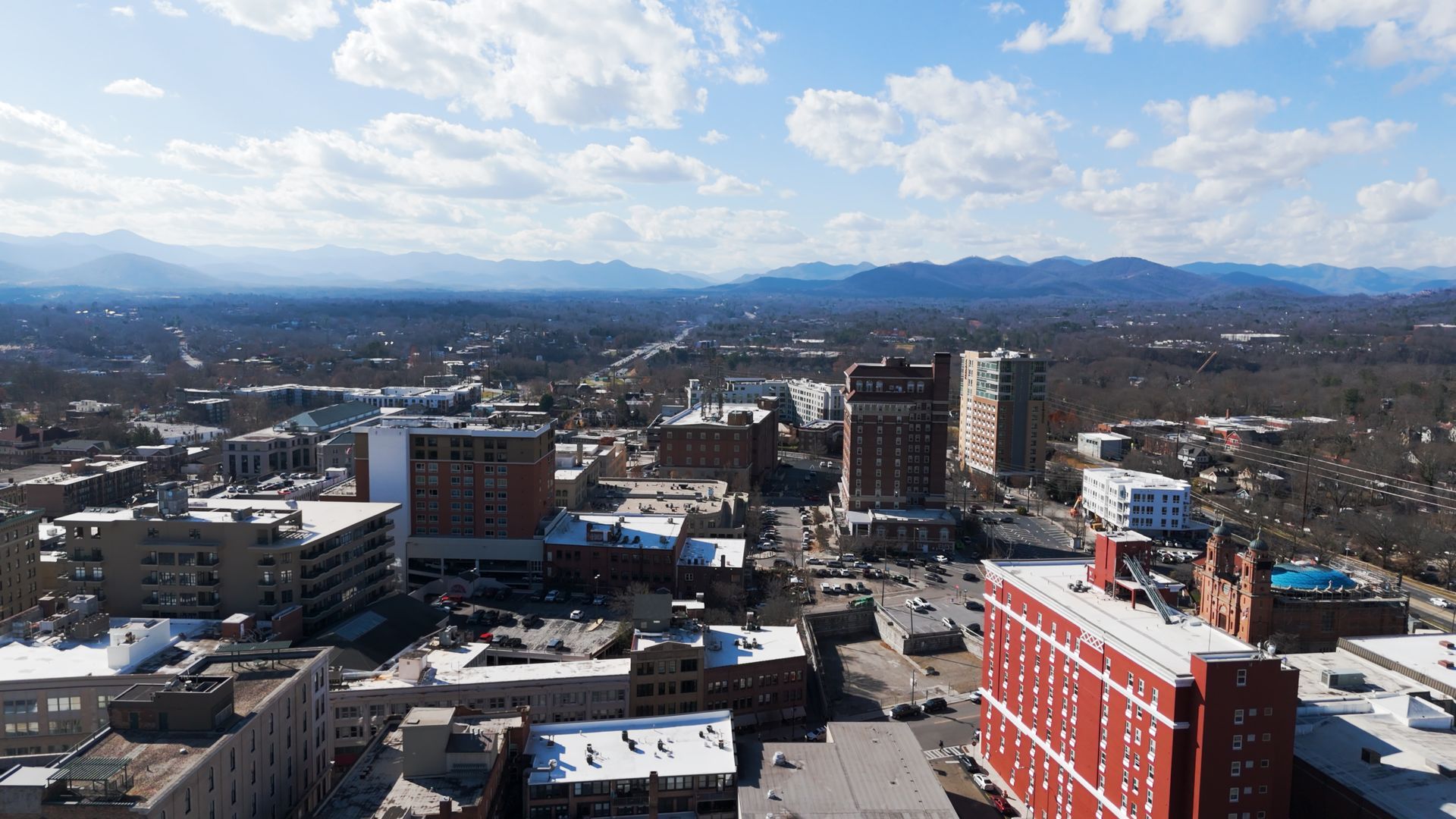 Cityscape with buildings and a red brick structure in the foreground, mountains in the distance under a clear blue sky.