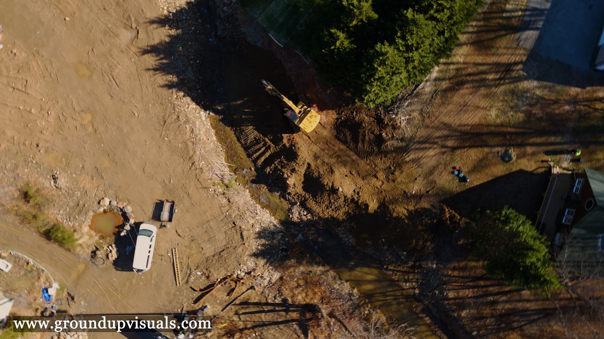 Aerial view of a dirt worksite with a white truck, equipment, and trees casting long shadows.