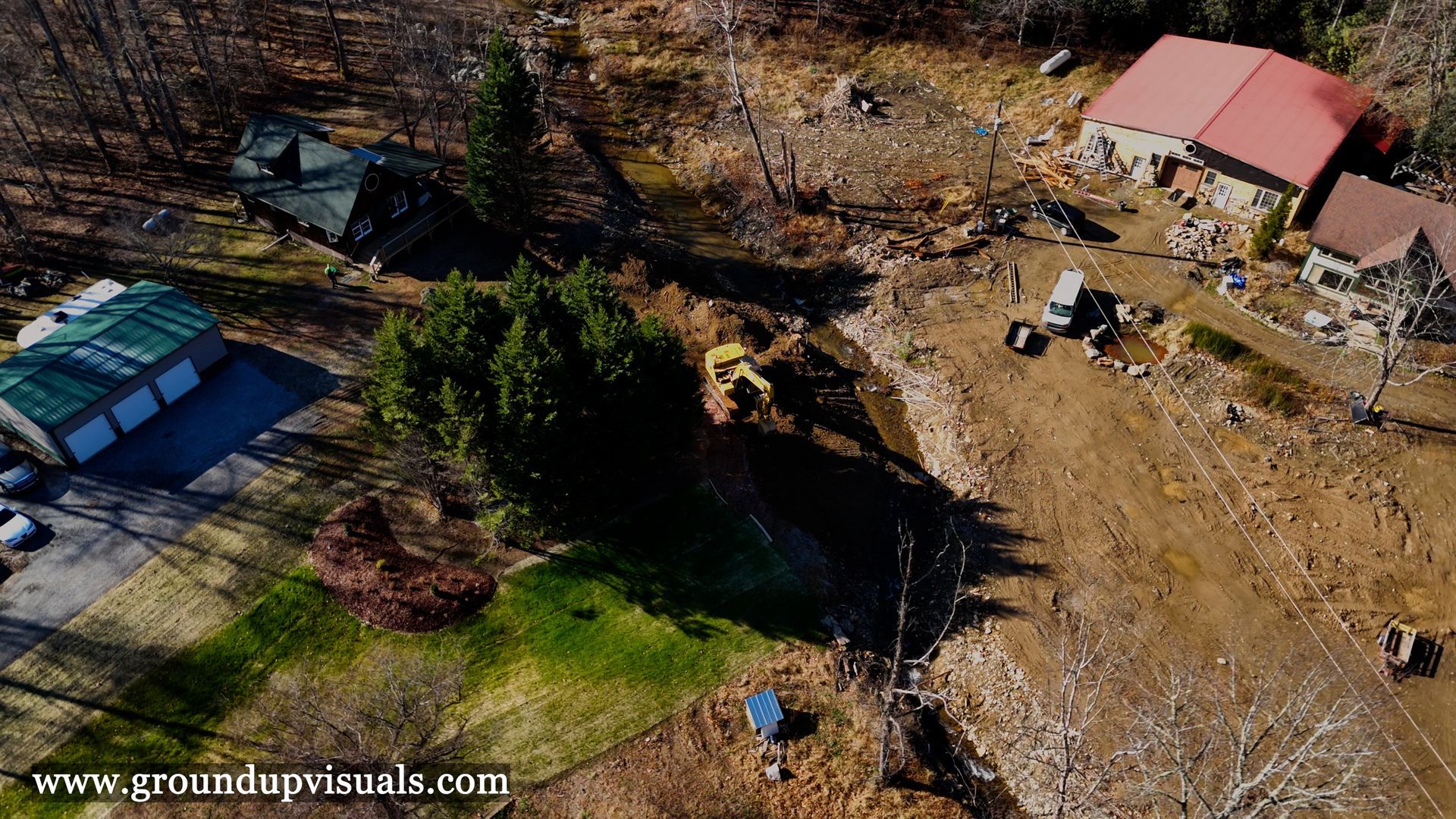 Aerial view of a rural yard with a red-roofed house, trees, scattered equipment, and open brown ground.