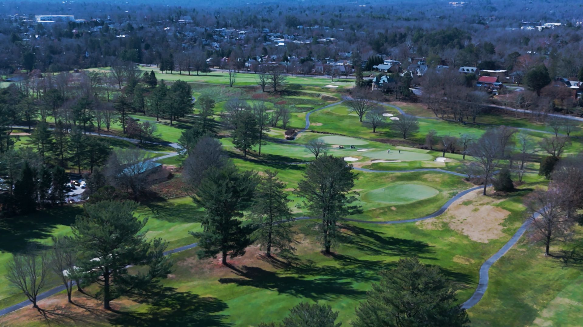 Aerial view of a green golf course with fairways, trees, sand bunkers, and nearby homes in the distance
