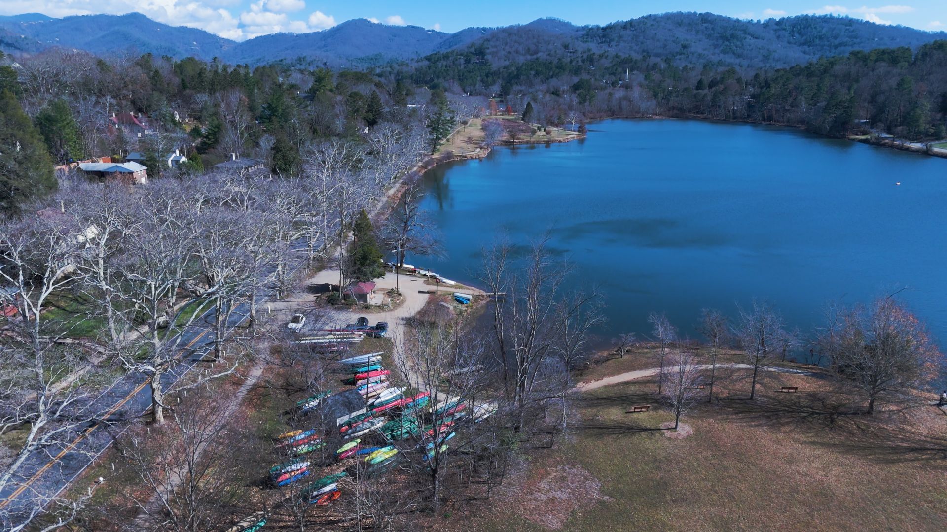 Aerial view of a blue lake beside a wooded shoreline and distant mountains, with a small riverside park.