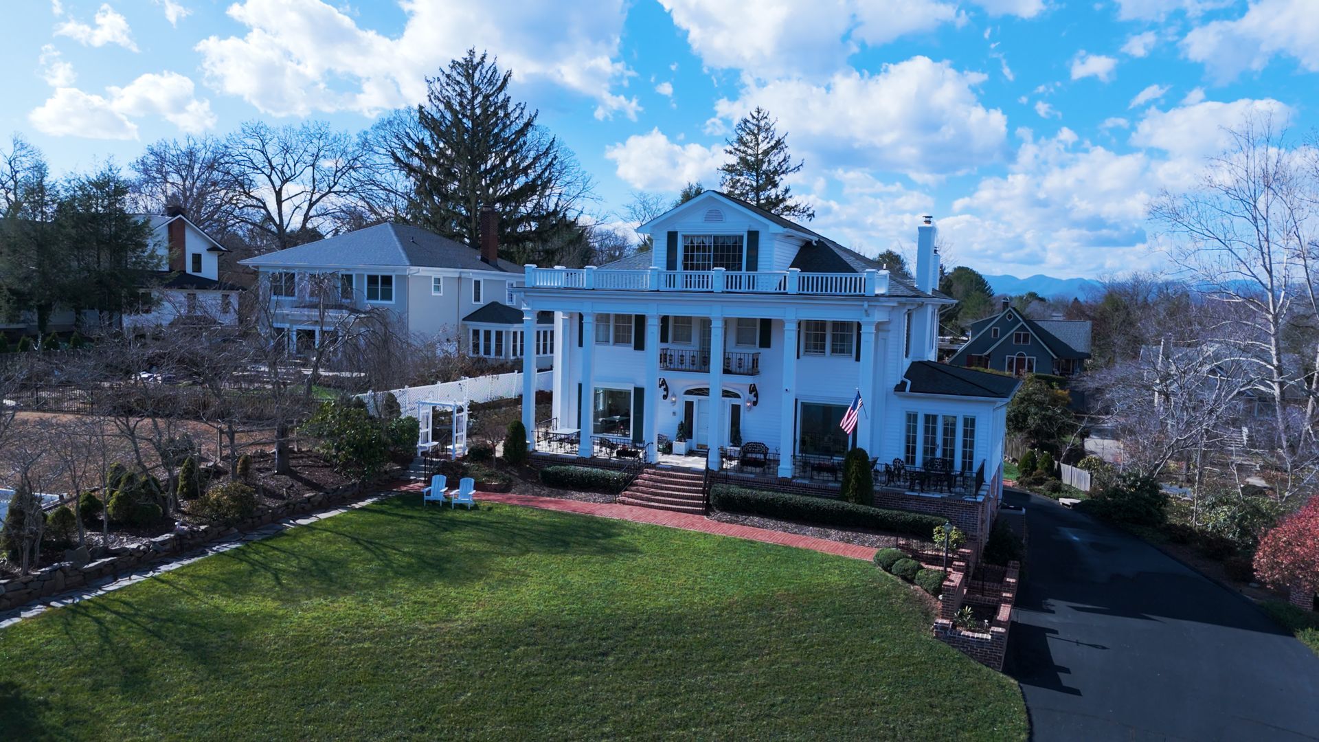 Large white colonial-style house with columns on a hill, overlooking a green lawn and winding driveway under a blue sky