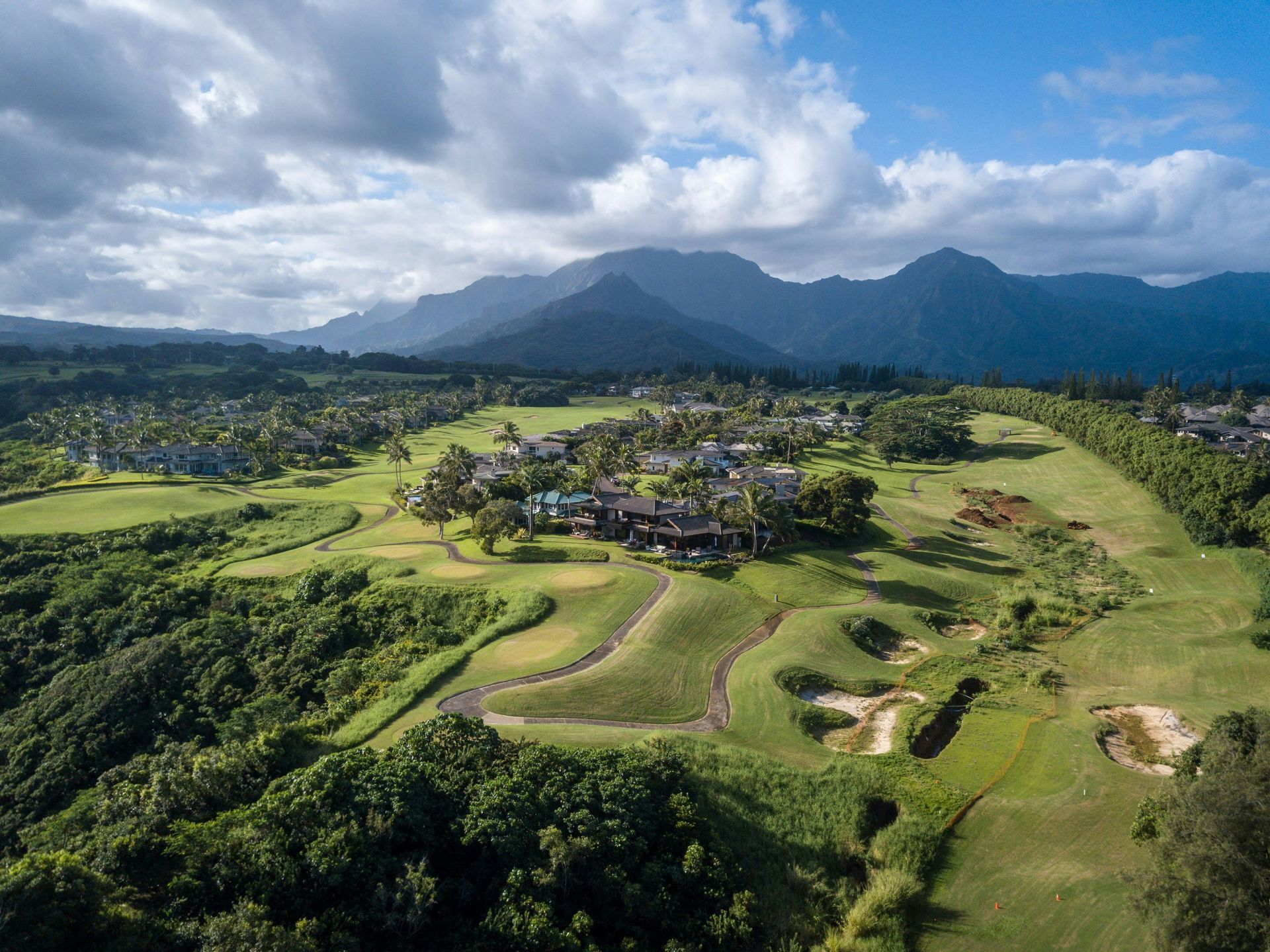 Green valley village with winding paths, lush fields, and mountains under a cloudy sky