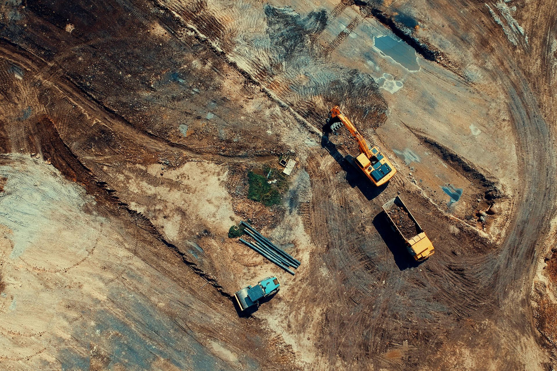 Aerial view of a construction site with two yellow excavators and a blue truck on bare ground.