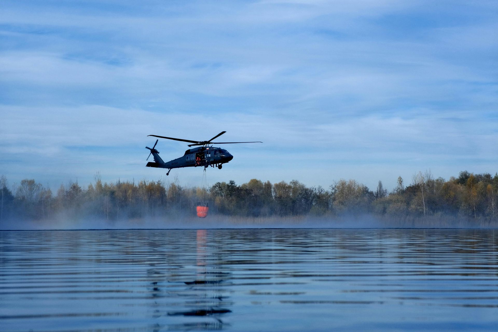 Black helicopter hovering low over calm water with mist and a forested shoreline at dawn
