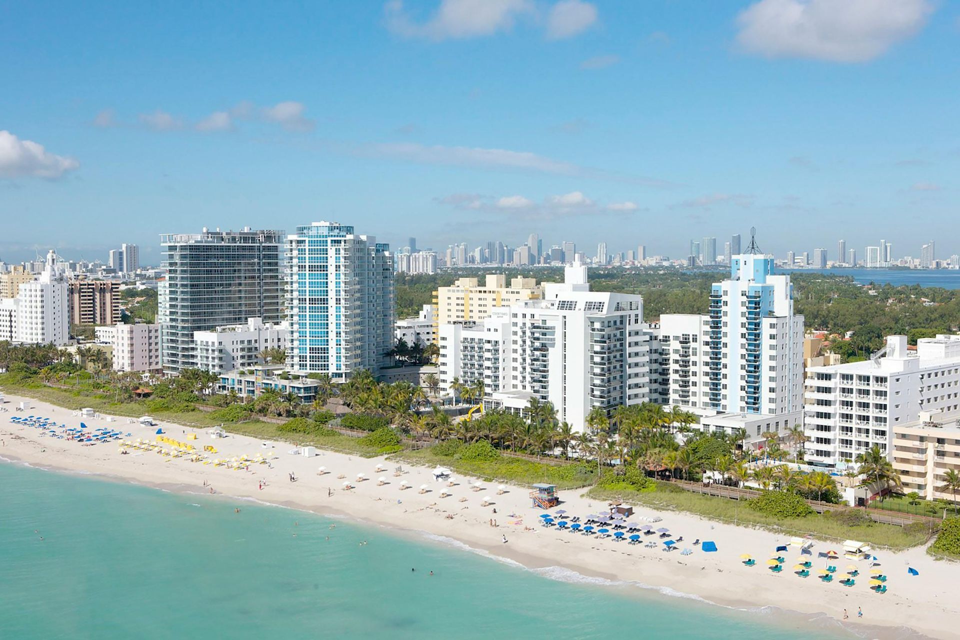 Sunny beach with turquoise water, white apartment buildings, and umbrellas along the shore under a blue sky