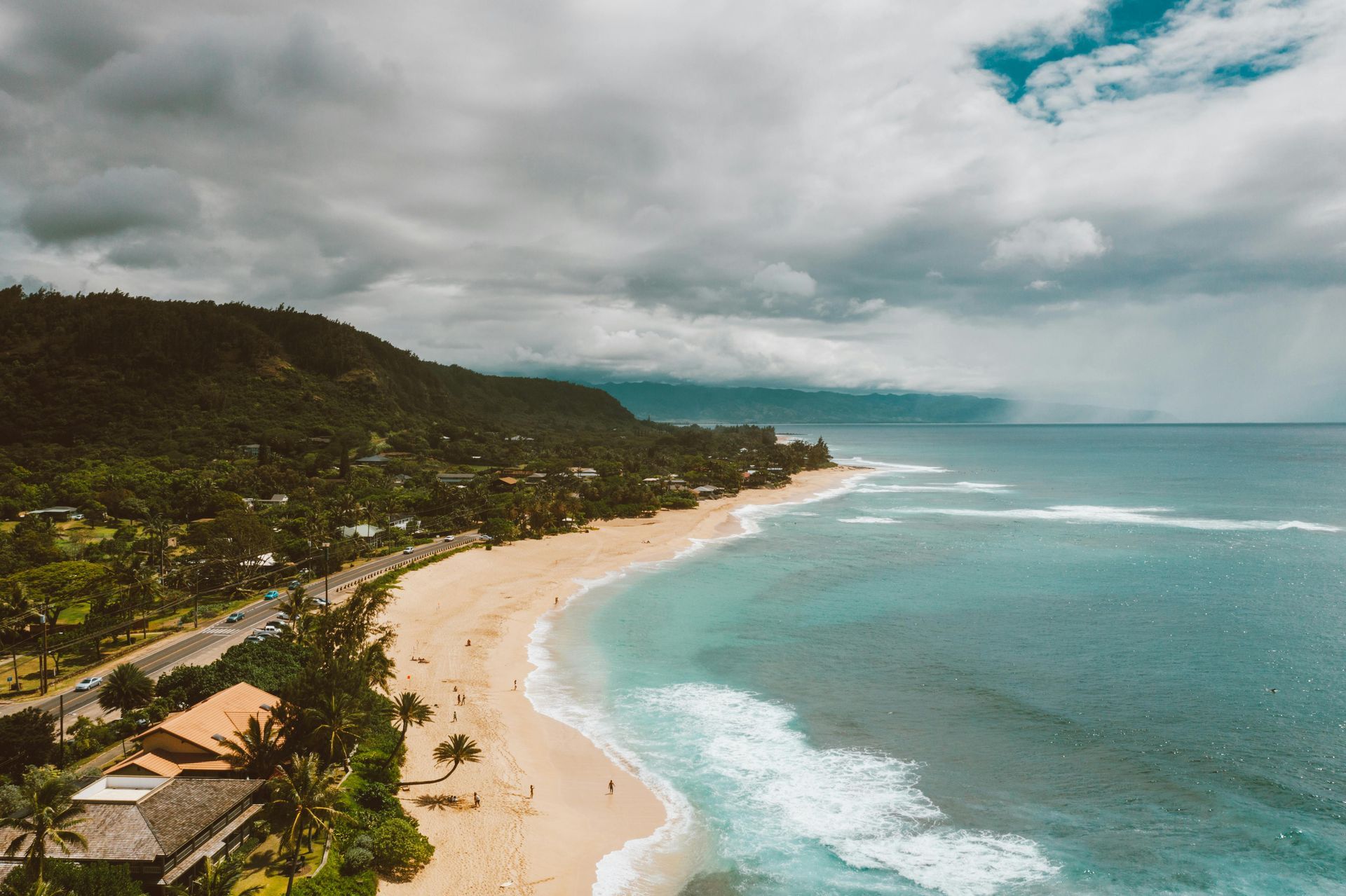Curved tropical beach with turquoise water, white sand, and lush green hills under a cloudy sky