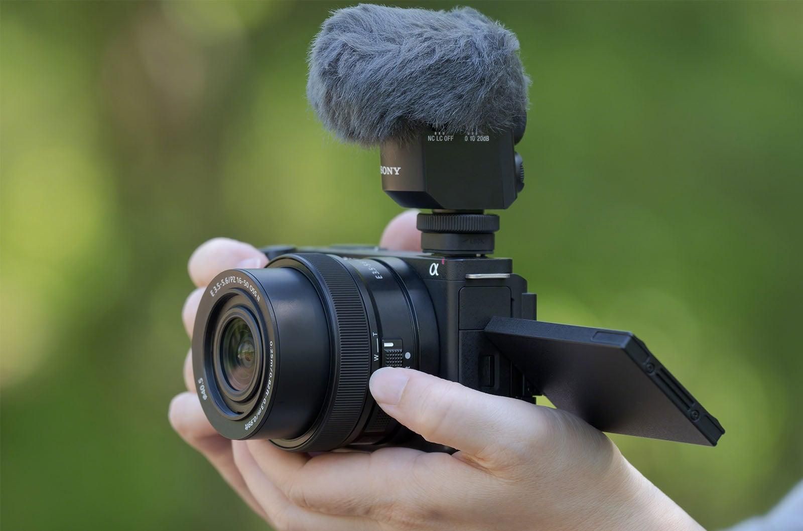 A black Sony mirrorless camera with a lens hood sits on a dark reflective surface against a vibrant yellow background.