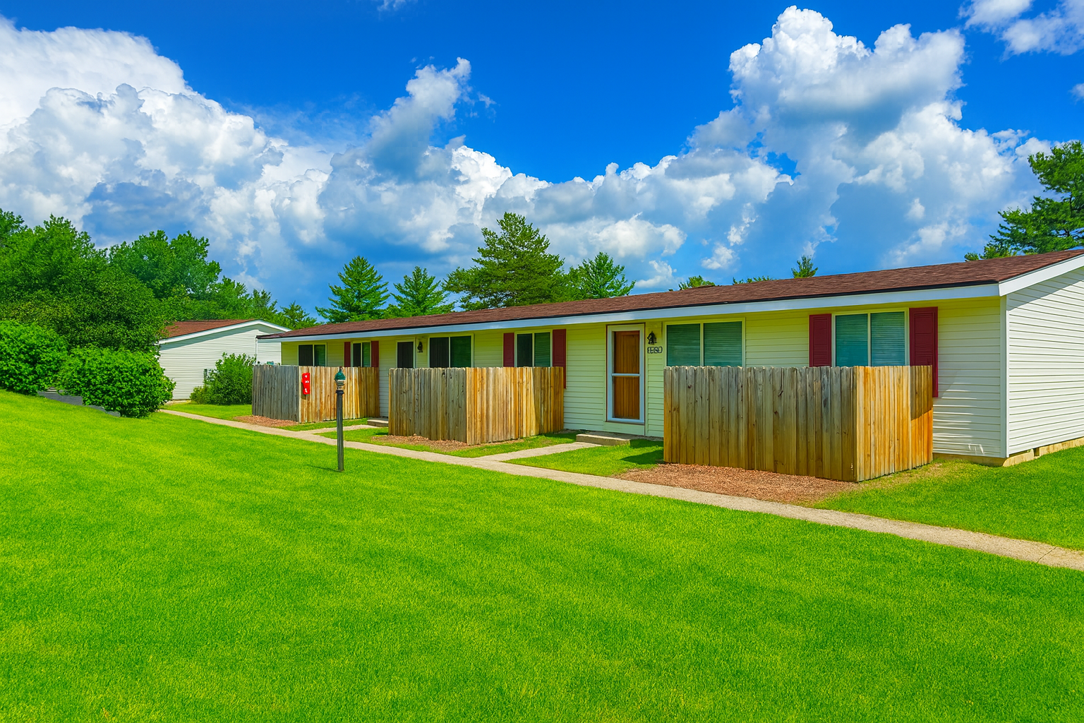 Row of white townhomes with brown roofs, wooden fences, and green lawn under a blue sky with clouds.