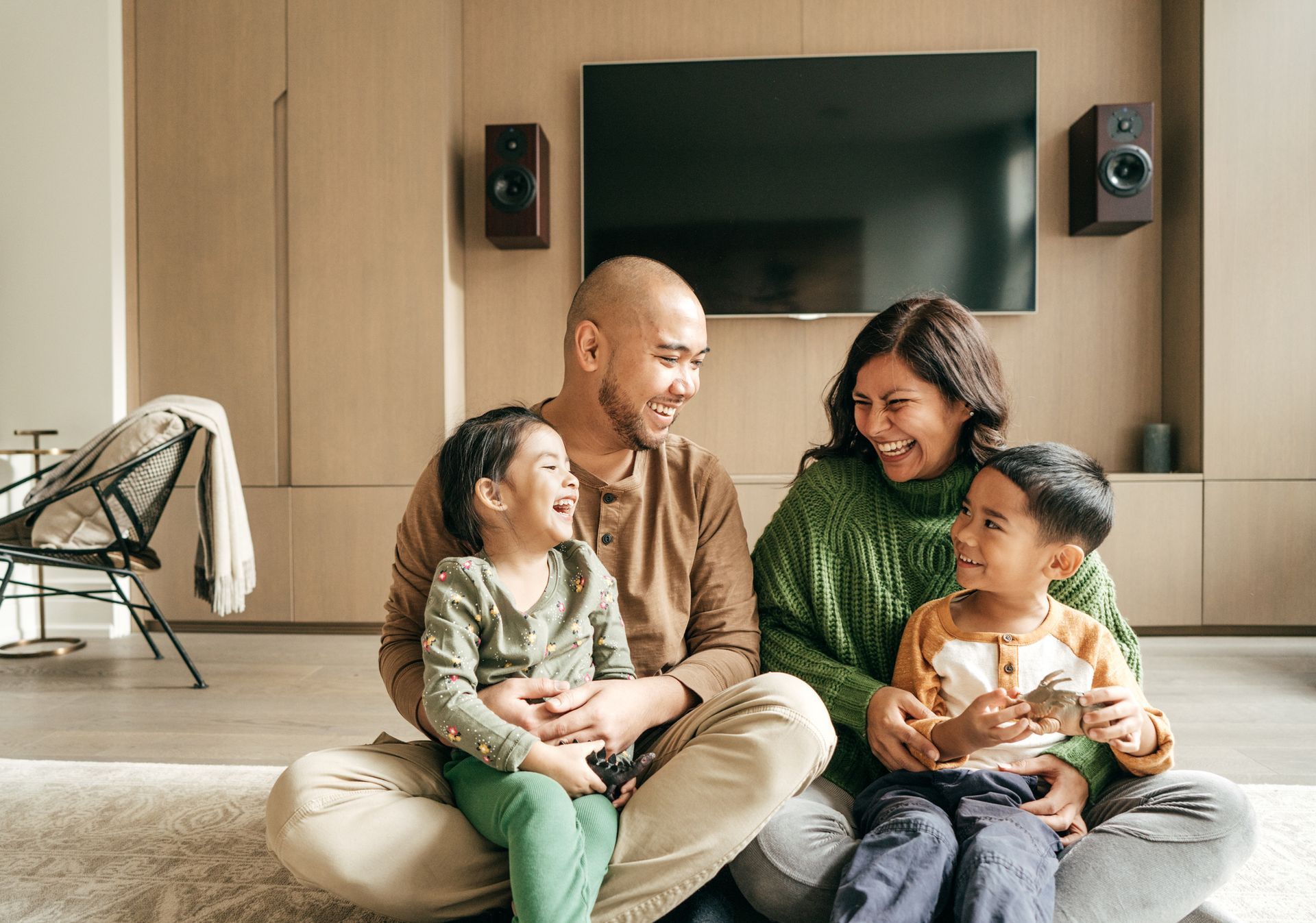 Family sitting on floor, smiling. Living room with TV, speakers, and chair visible.