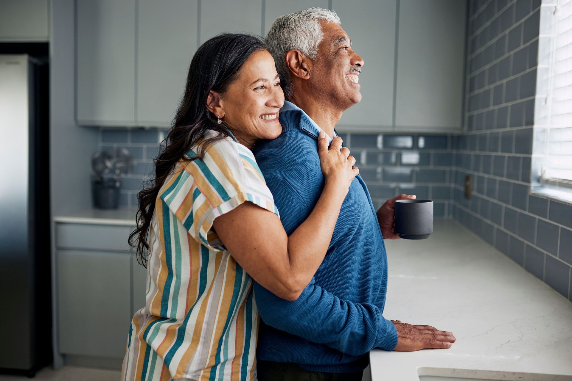Woman embraces man from behind in a kitchen, both looking out the window, smiling.