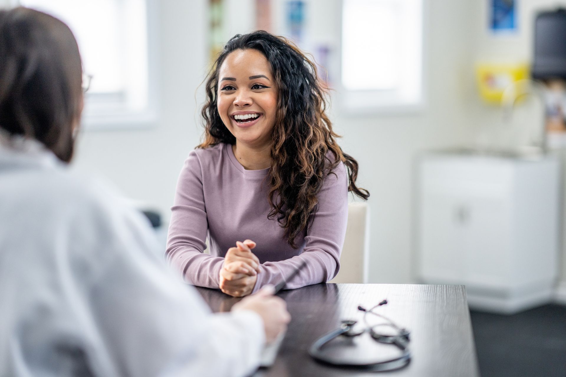 Woman smiling in a medical office, facing a doctor, seated at a desk.
