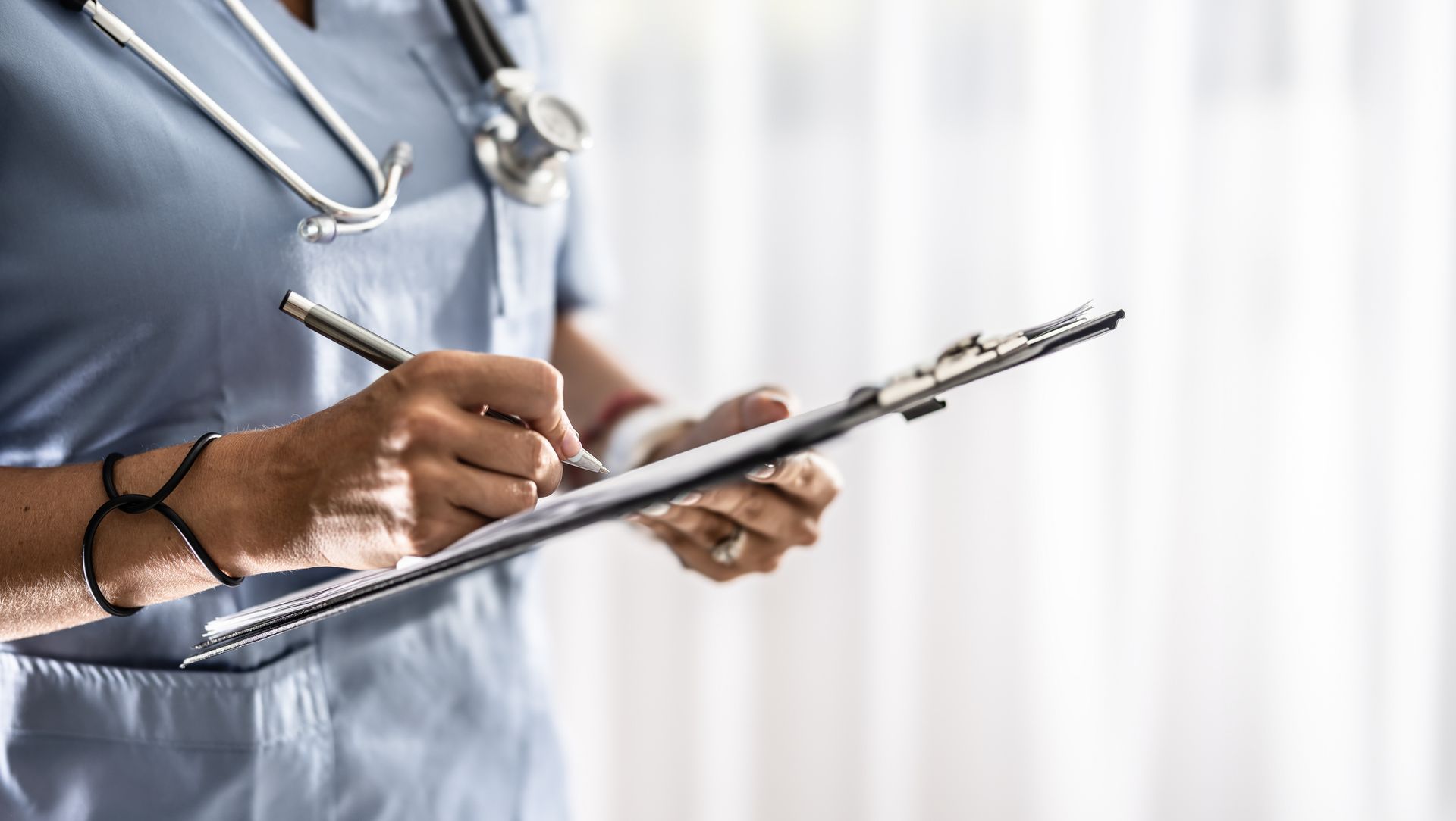 Nurse in blue scrubs writes on a clipboard, stethoscope around neck.