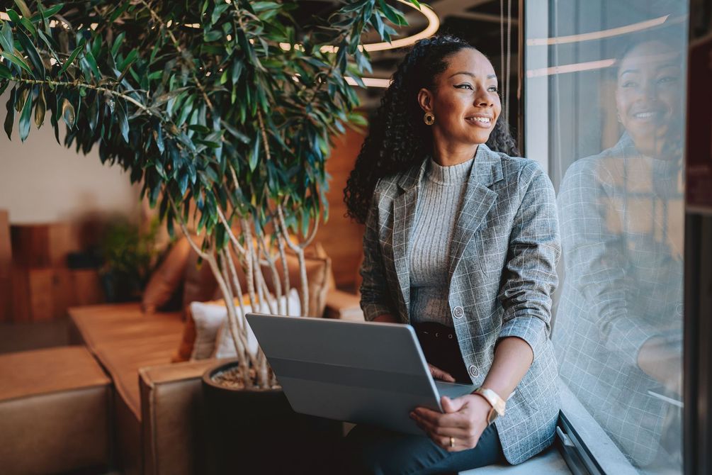 Woman holding laptop, looking out window, smiling. Sitting near a plant in an office setting.