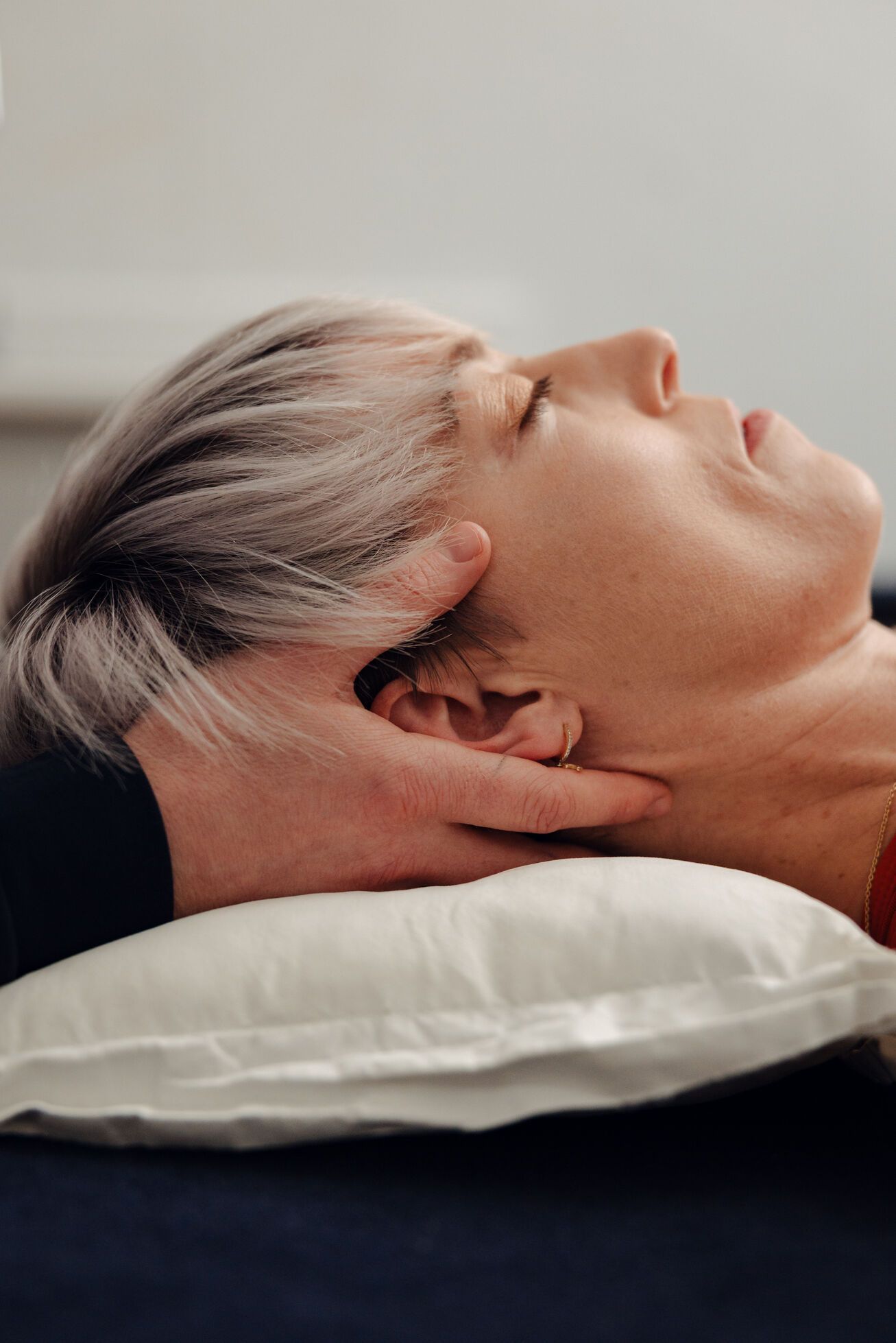 Woman with short silver hair lies on pillow and receives cranial osteopathy with a practitioner holding the back of her head.
